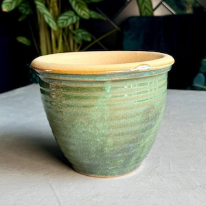 May include: A green ceramic plant pot with a textured surface and a light tan rim. The pot has a wide opening and a slightly tapered shape. The pot is empty and ready for planting. The pot is sitting on a grey surface.