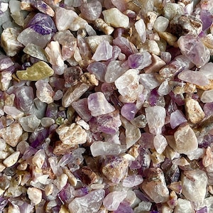 May include: A pile of small, purple and clear amethyst crystals on a grey background.