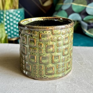 May include: A small, cylindrical ceramic planter with a textured, square pattern in shades of green and gold. The interior is dark, and the planter sits on a light grey surface. The design gives a vintage or retro feel, suitable for small plants or succulents.