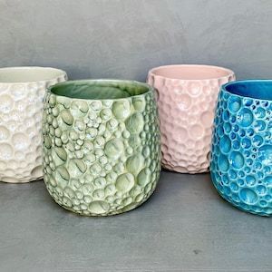 May include: Four ceramic pots in cream, green, pink, and blue. Each has a textured, bubble-like surface. The pots are cylindrical with a slightly wider top. The colours are soft and the overall aesthetic is modern.