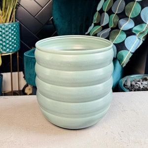 May include: A light green ceramic planter with a ribbed design. The planter is sitting on a white surface.