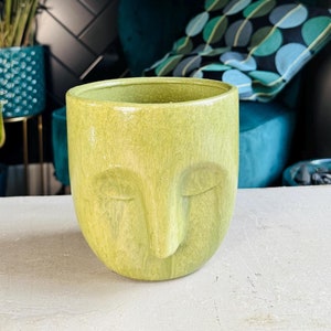 May include: A green ceramic planter shaped like a face with closed eyes and a prominent nose. The planter is sitting on a white surface.