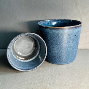 May include: Two blue ceramic mugs, one slightly larger than the other, with a speckled glaze. The mugs are sitting on a white surface.