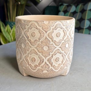 May include: A beige ceramic plant pot with a detailed, raised geometric pattern. The pot has a cylindrical shape with a slightly flared top and small feet. The neutral colour and textured design give it a rustic, yet elegant appearance, suitable for various home decor styles.