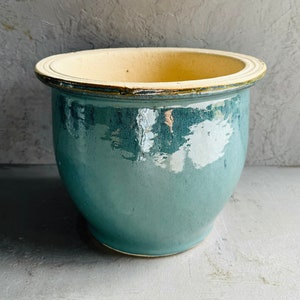 May include: A teal ceramic plant pot with a glossy finish. The pot has a wide, rounded shape with a cream-coloured rim. The interior is also cream-coloured. Suitable for indoor or outdoor use.