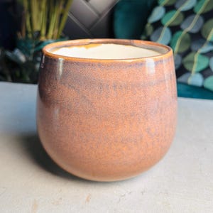 May include: A rounded, copper-toned ceramic plant pot with a speckled finish. The pot has a wide opening and is filled with a white substance. A green plant and teal armchair are visible in the background.