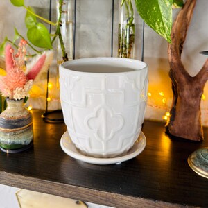 May include: A white ceramic plant pot with a decorative embossed pattern, sitting on a matching saucer. The pot is on a dark wooden shelf, with other decorative items and plants in the background. The overall theme is home decor.