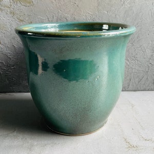 May include: A teal green ceramic planter with a slightly flared rim. The planter has a glossy finish and is sitting on a white surface.