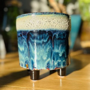 May include: A ceramic plant pot with a distinctive glaze. The top rim is speckled beige, flowing into a vibrant blue and turquoise with a dripped effect. It stands on three short, dark brown legs, adding a touch of elegance to this decorative piece.