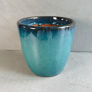 May include: A turquoise ceramic planter with a glossy finish. The planter has a wide opening and a rounded shape, with a dark blue rim and a lighter turquoise colour that fades down the sides. The interior is a reddish-brown colour.