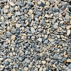 May include: Close-up of a pile of small, irregularly shaped rocks. The rocks are primarily shades of grey, blue-grey, and tan, creating a natural, textured appearance. The varying sizes and colours suggest a gravel or aggregate material.