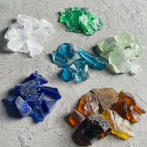 May include: Assortment of colourful crushed glass pieces in various shades of blue, green, brown, and white. The glass pieces are arranged in separate piles on a grey textured surface.