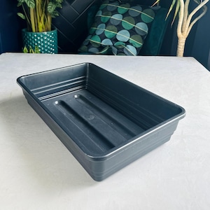 May include: A dark grey rectangular plant tray with a ridged interior. The tray is made of plastic and is designed for holding plants. The tray is on a white surface.