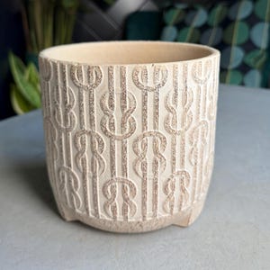 May include: A cream-coloured ceramic planter with a repeating pattern of interlocking shapes. The cylindrical pot has a textured surface and sits on three small feet. The interior is a lighter shade of cream.