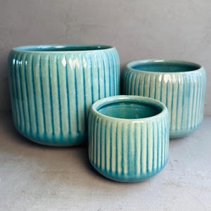 May include: Three turquoise ceramic plant pots of different sizes. Each pot features a ribbed, vertical design. The pots are arranged together, showing their different heights and widths. The colour is a vibrant teal.