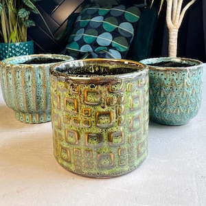 May include: Three ceramic plant pots in shades of green and teal. The pots have textured surfaces with geometric and patterned designs. The pots are empty and ready for plants. The pots are of varying sizes and shapes.