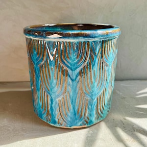 May include: A ceramic plant pot with a turquoise and bronze feather design. The cylindrical pot has a rounded rim and a textured surface. The colour palette includes shades of blue, brown, and gold. This decorative pot is suitable for indoor or outdoor use.