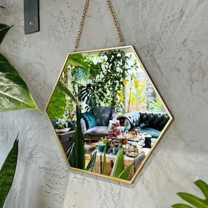 May include: A gold hexagonal mirror with a chain hanging from the top. The mirror is reflecting a room with plants and furniture.
