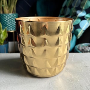 May include: A gold-coloured planter with a geometric pattern. The planter is made of a textured material and has a round shape.