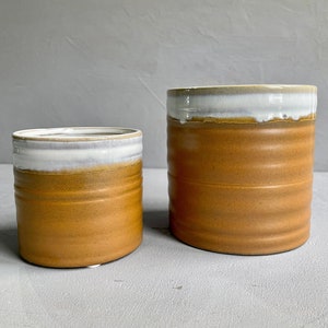 May include: Two ceramic pots with a white rim and a brown glaze. The smaller pot is in front of the larger pot.
