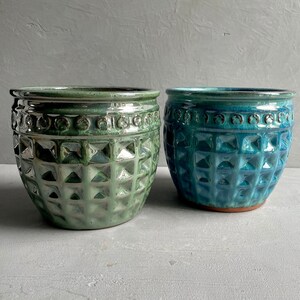 May include: Two ceramic plant pots, one teal and the other green. Both have a textured, geometric pattern and a decorative band near the rim. The pots are round and have a glossy finish. The pots are suitable for indoor or outdoor use.