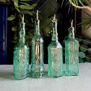 May include: Four decorative glass bottles with silver spouts. The bottles are various shapes and sizes, with embossed floral and geometric patterns. The glass has a light green tint. The bottles are arranged on a light grey surface.
