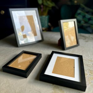 May include: Several black picture frames of different sizes, some with white mounts. The frames are empty, ready for photos or artwork. They are arranged on a light-coloured surface, with a blurred background.
