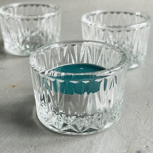 May include: Three clear glass votive candle holders with a diamond-cut pattern. One holder contains a teal-coloured candle. The holders are round with a thick base, suitable for home decor or special events.