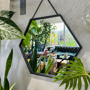 May include: A black hexagonal mirror with a chain hanging from the top. The mirror is reflecting a room with plants and furniture.