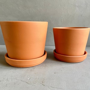 May include: Two terracotta plant pots with matching saucers. The larger pot is taller and wider than the smaller pot.