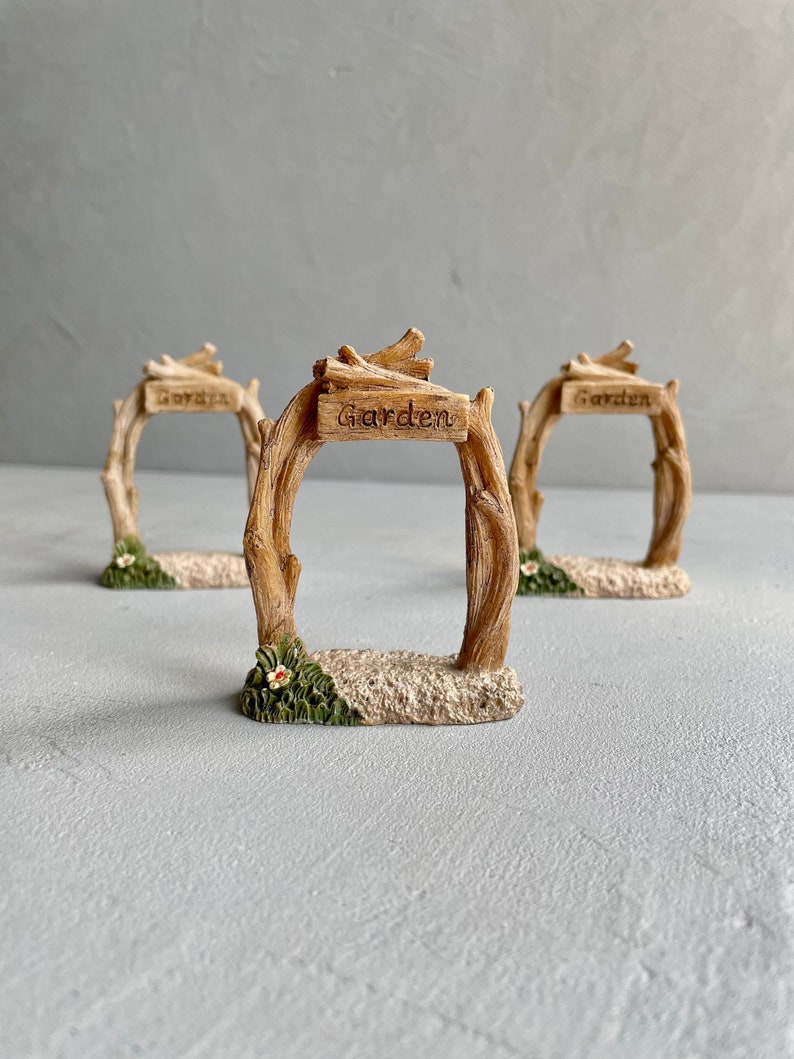 May include: Three miniature wooden garden arches with the word "Garden" inscribed on each. The arches are made of brown twigs and have a sandy base with green grass and a white flower.