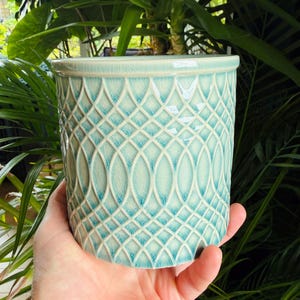 May include: A cylindrical ceramic plant pot featuring a light turquoise and cream glaze with a distinct crackle finish. The exterior surface is decorated with a repeating embossed lattice pattern consisting of intersecting oval shapes.