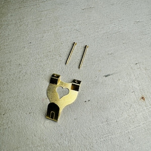 May include: A gold-colored metal picture hook with a heart-shaped cutout. Two gold-colored nails are included.