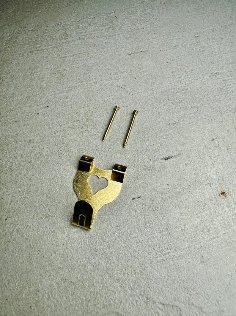 May include: A gold-colored metal picture hook with a heart-shaped cutout. Two gold-colored nails are included.
