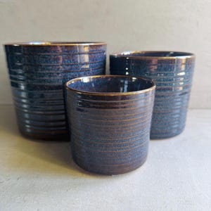 May include: Three ceramic plant pots in different sizes. The pots are a deep blue with a textured, horizontal ribbed design. The rims have a gold-coloured trim. Suitable for indoor or outdoor use, these pots are ideal for plants.
