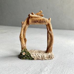 May include: A miniature wooden garden archway with a sign that reads "Garden". The archway is made of brown branches and has a small flower at the base.