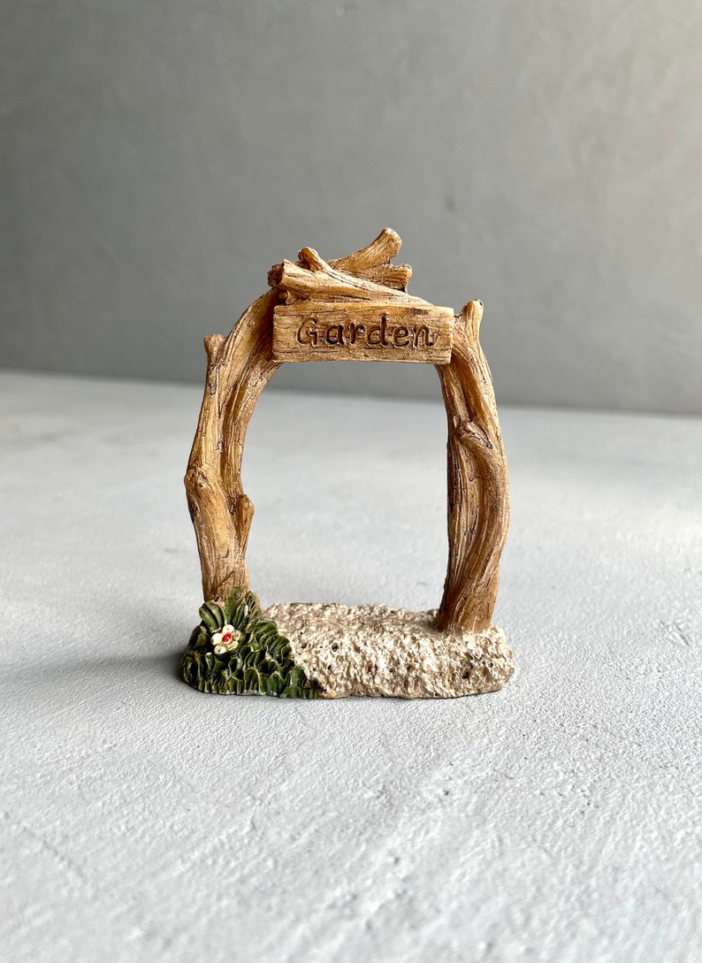 May include: A miniature wooden garden archway with a sign that reads "Garden". The archway is made of brown branches and has a small flower at the base.