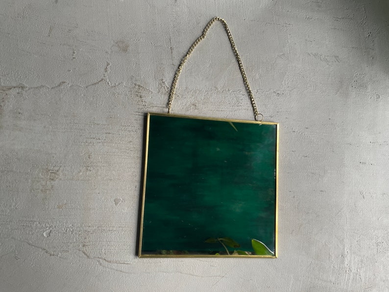 May include: A gold-colored metal picture frame with a dark green background. The frame has a chain attached to the top for hanging.