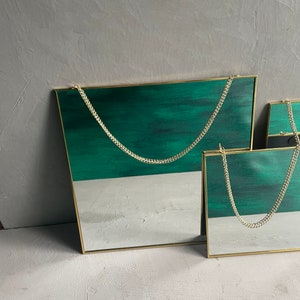 May include: Two square mirrors with gold frames and a green background. The mirrors are hanging from a gold chain.