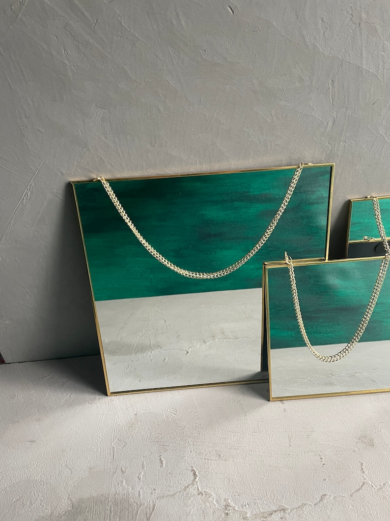 May include: Two square mirrors with gold frames and a green background. The mirrors are hanging from a gold chain.