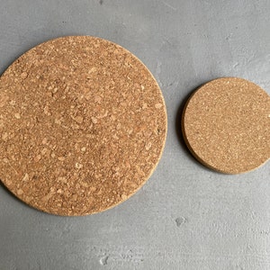 May include: Two round cork trivets, one larger than the other, on a grey surface. The trivets are made of natural cork and have a rough texture.