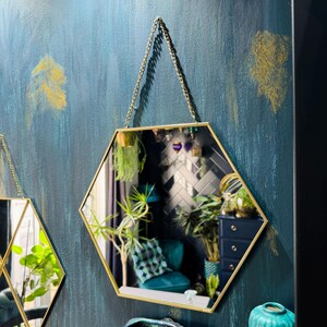 May include: Two gold hexagonal mirrors with a chain hanging from the top. The mirrors are hanging on a teal blue wall with gold accents. A teal blue vase is on the bottom right corner of the image.