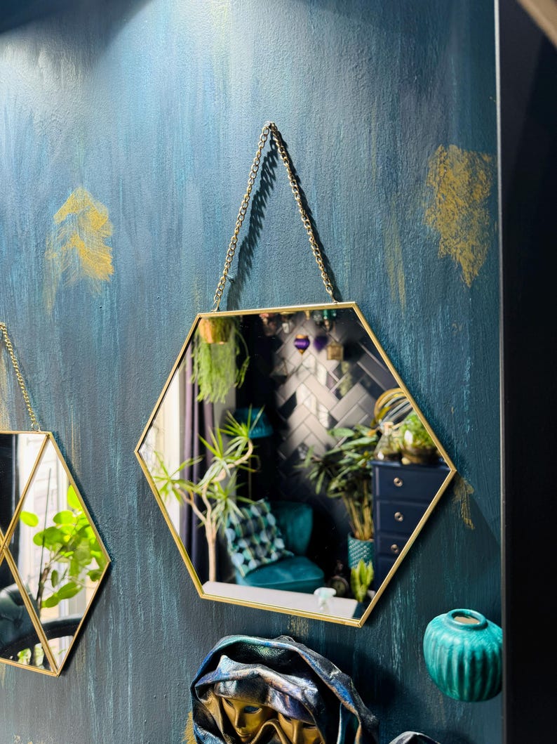May include: Two gold hexagonal mirrors with a chain hanging from the top. The mirrors are hanging on a teal blue wall with gold accents. A teal blue vase is on the bottom right corner of the image.