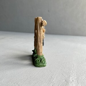 Pu&ograve; includere: Un piccolo recinto in legno in miniatura con una base verde, che presenta una piccola figura di una persona appoggiata al recinto.