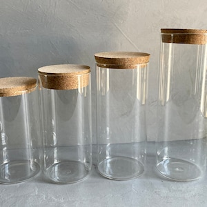 May include: Four clear glass jars with cork lids of varying sizes. The jars are empty and arranged in a row from largest to smallest.