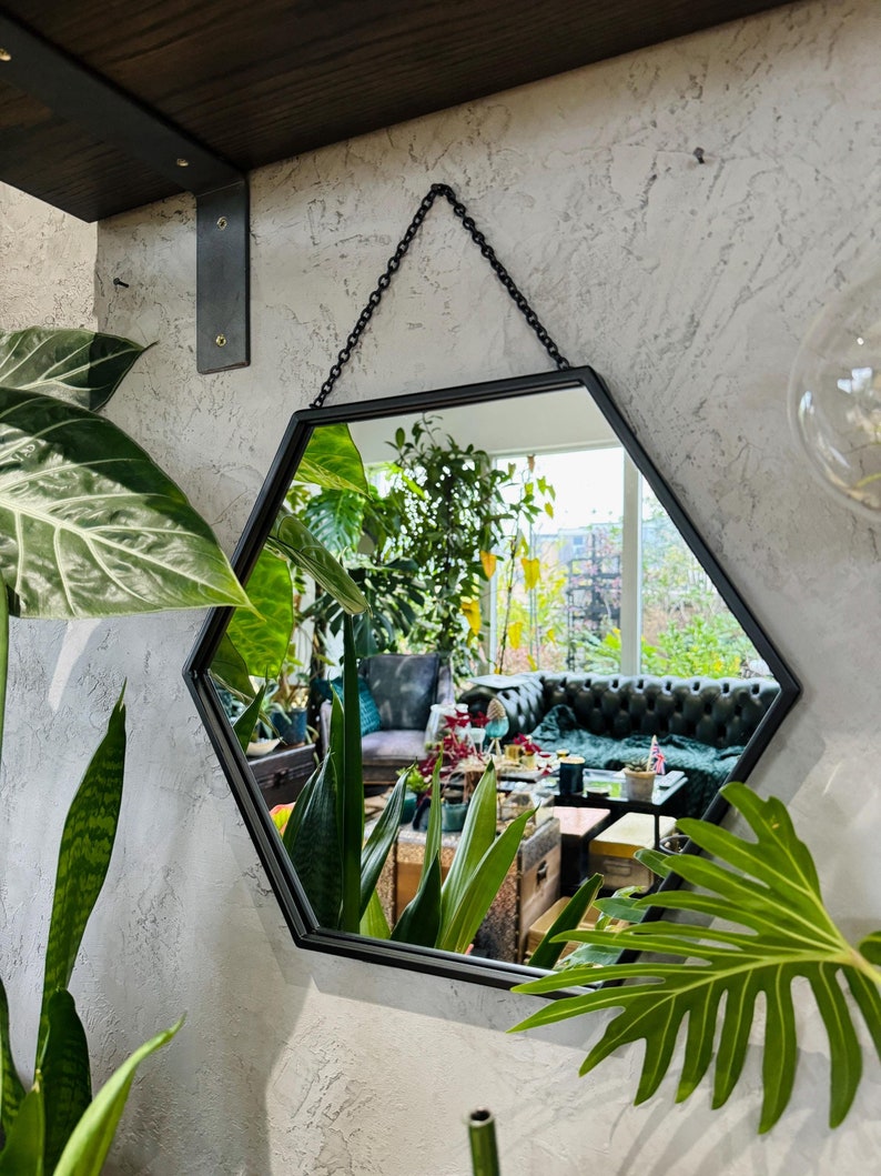 May include: A black hexagonal mirror with a chain hanging from the top. The mirror is reflecting a room with plants and furniture.