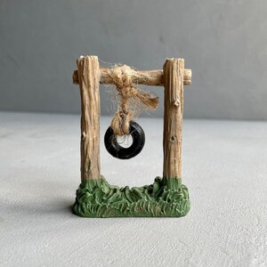 Pu&ograve; includere: Un'altalena in legno in miniatura con un pneumatico nero appeso. L'altalena &egrave; su una base verde che sembra erba.
