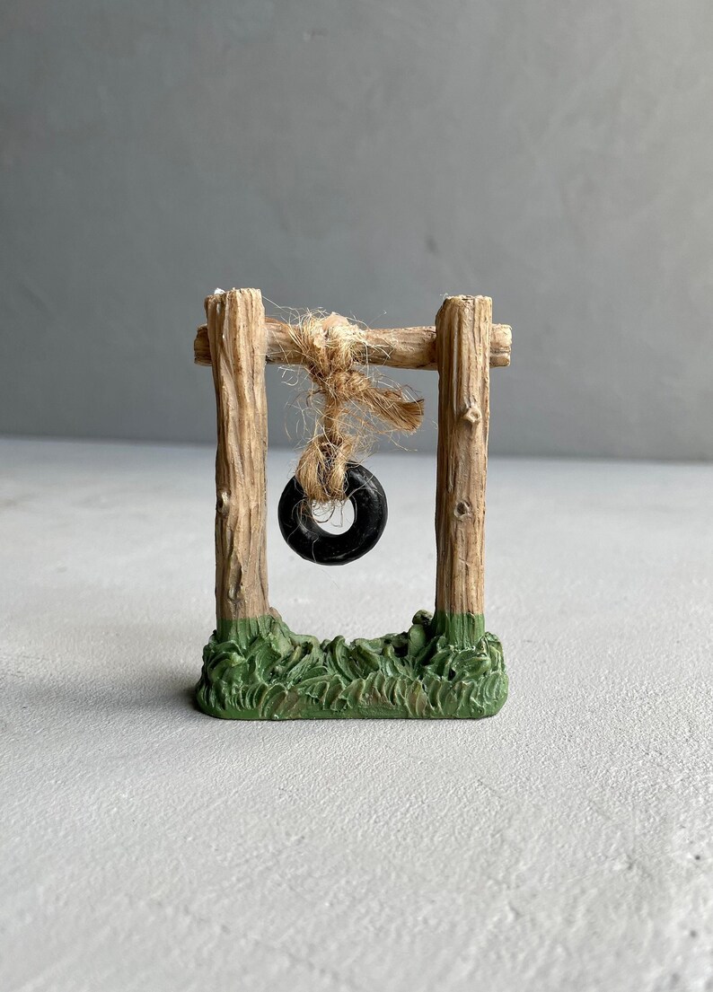 Pu&ograve; includere: Un'altalena in legno in miniatura con un pneumatico nero appeso. L'altalena &egrave; su una base verde che sembra erba.