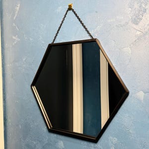 May include: A black hexagonal mirror with a chain hanging from a gold hook on a blue textured wall.