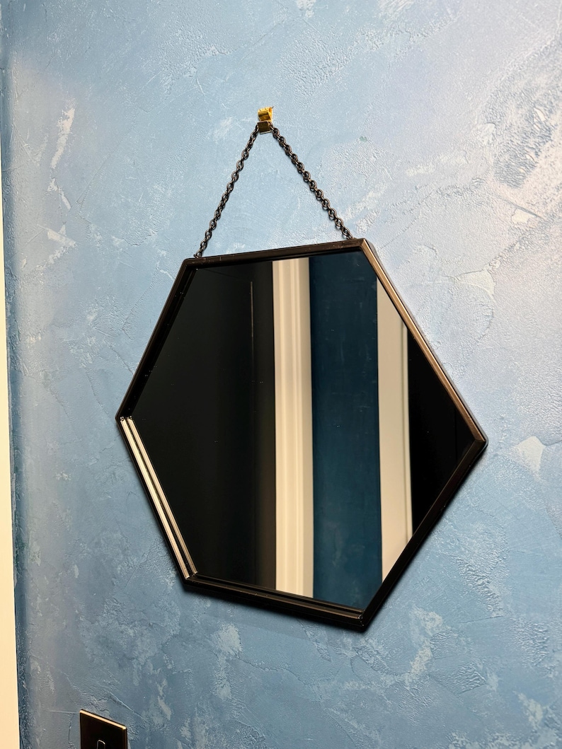 May include: A black hexagonal mirror with a chain hanging from a gold hook on a blue textured wall.